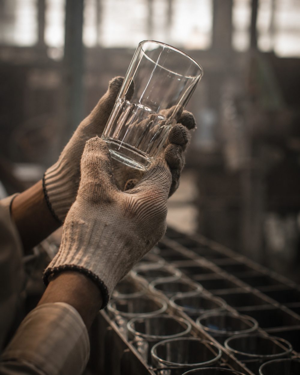 A craftsman's gloved hands inspecting a clear glass tumbler against window light at Glastic's factory in Firozabad