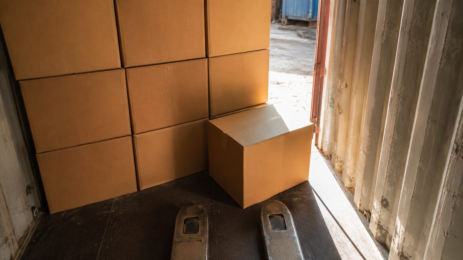 Detail inside a part-loaded shipping container, courses of kraft master cartons and a pallet truck's forks